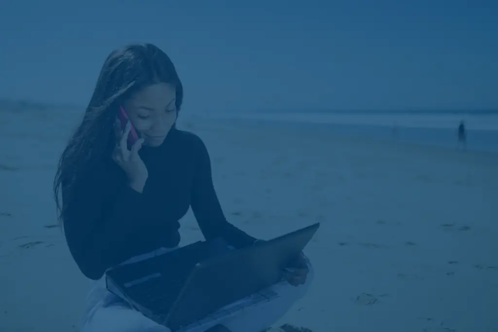 Young woman sitting on a beach while working on a laptop and making a phone call